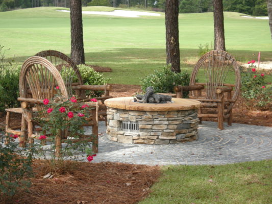 round stone gas firepit on a round patio with willow wooden furniture in a garden by a golf course