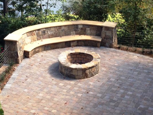 round stone firepit and seating wall on a brick and stone patio in a backyard garden
