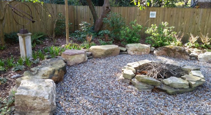 rustic round stone firepit on a gravel patio in a backyard garden with garden art