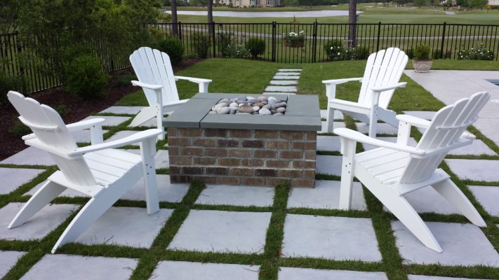 square stone fire-pit on a stone patio with four Adirondack chairs in a backyard garden