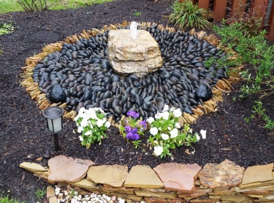 chunky stone didappearing fountain bubbling water into a ring of black mexican beach pebbles and mulch by flowers at stone garden