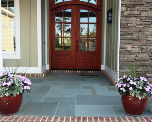 stone patio porch at the double front door entrance of a home flanked by colorful flowers in stone pots