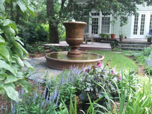 large stone urn fountain bubbling water into a round pool in a lush backyard garden