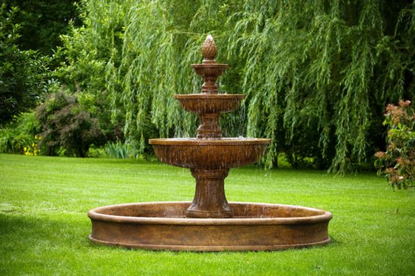 large stone triple-tiered fountain bubbling water into a round pool in a lush, green backyard garden