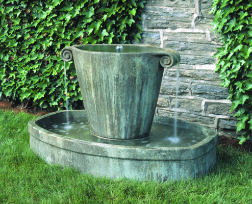 green, stone, urn garden fountain with water spilling over into an oblong basin in a garden by a stone wall