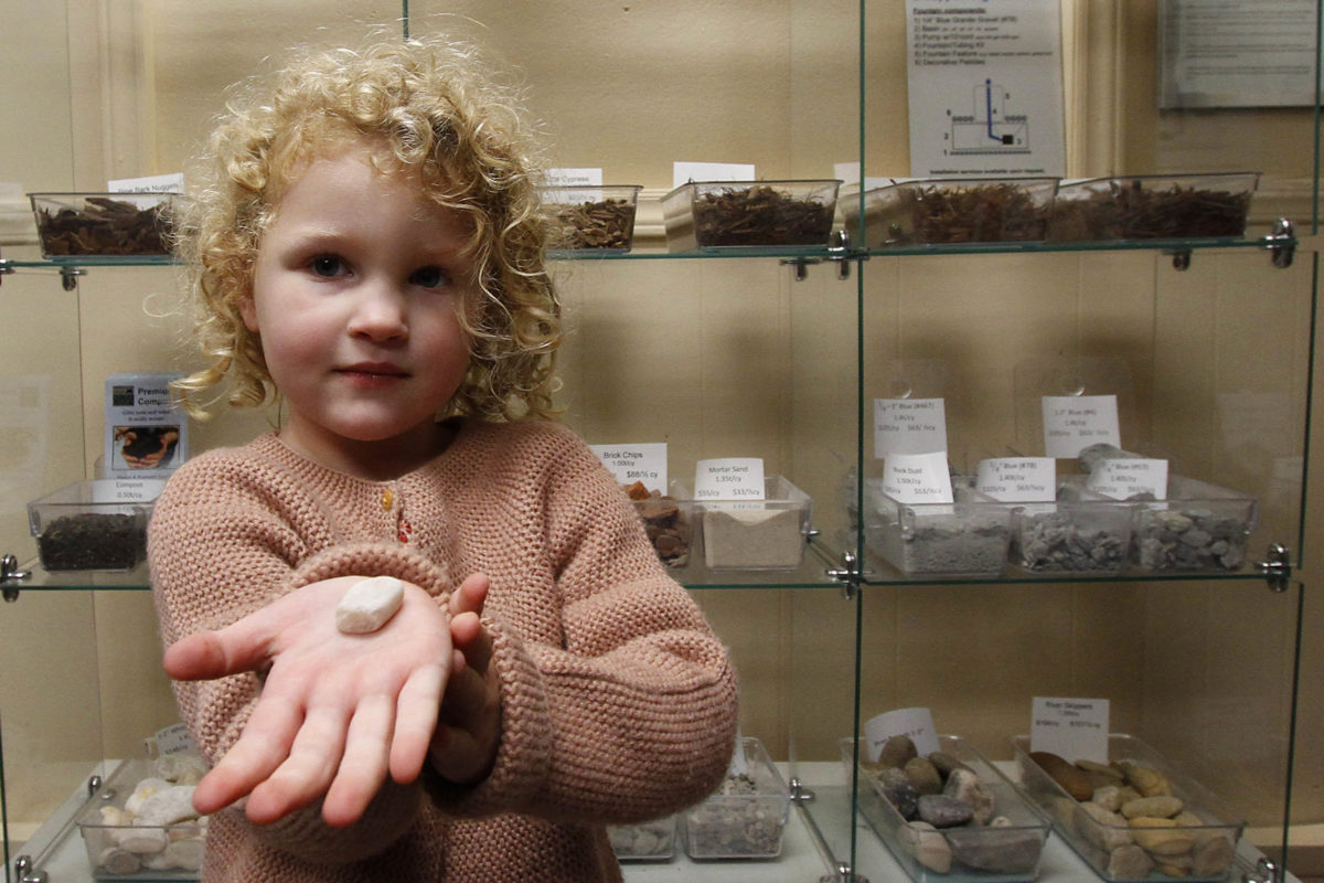 little, curly-haired girl holding rock in her palm by display case at stone garden