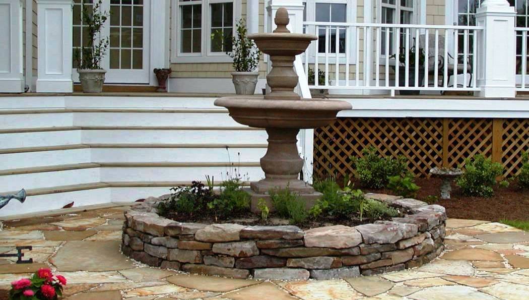 two-tiered stone fountain in a round, stone garden sitting on a flagstone patio in the backyard of a home