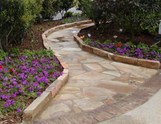 stone blocks edge a flagstone pathway through a garden of purple flowers and green trees