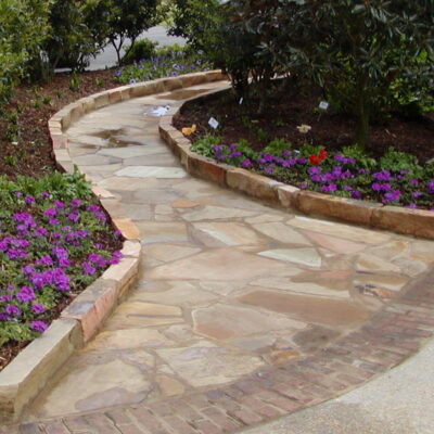 stone blocks edge a flagstone pathway through a garden of purple flowers and green trees