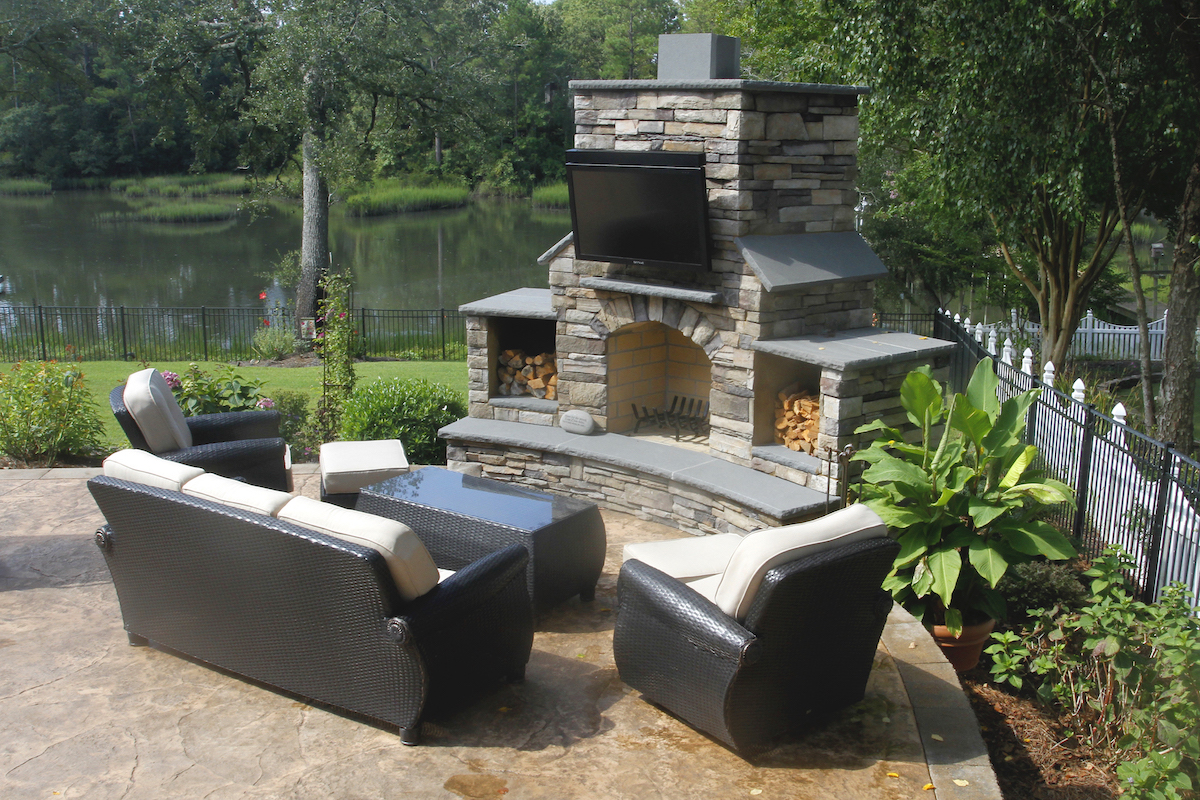 outdoor living room featuring a stone fireplace with television on a patio overlooking a pond