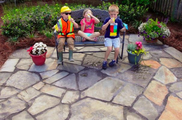 stone garden patio three children drinking juice boxes sitting on a metal bench in a backyard