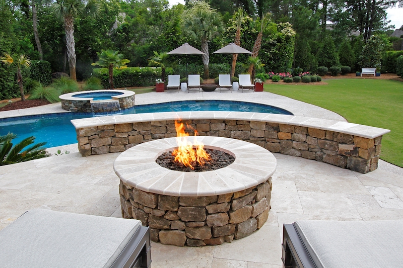 stone gas firepit on a travertine patio overlooking a pool and spa by a green lawn in a backyard garden