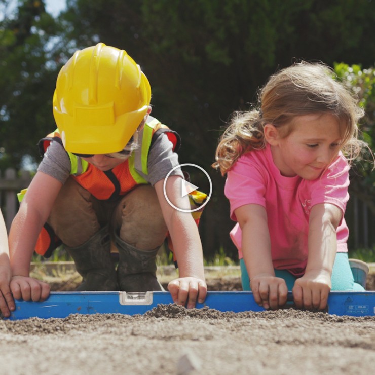 boy in a yellow hard hat and girl in a pink shirt using a level on sand