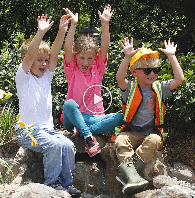 three happy children sitting on an outdoor backyard waterfall with their hands in the air
